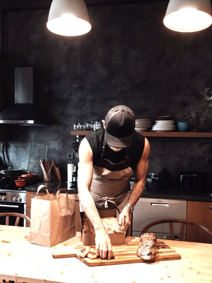 A baker packing an order into a paper bag on the counter of a small kitchen.